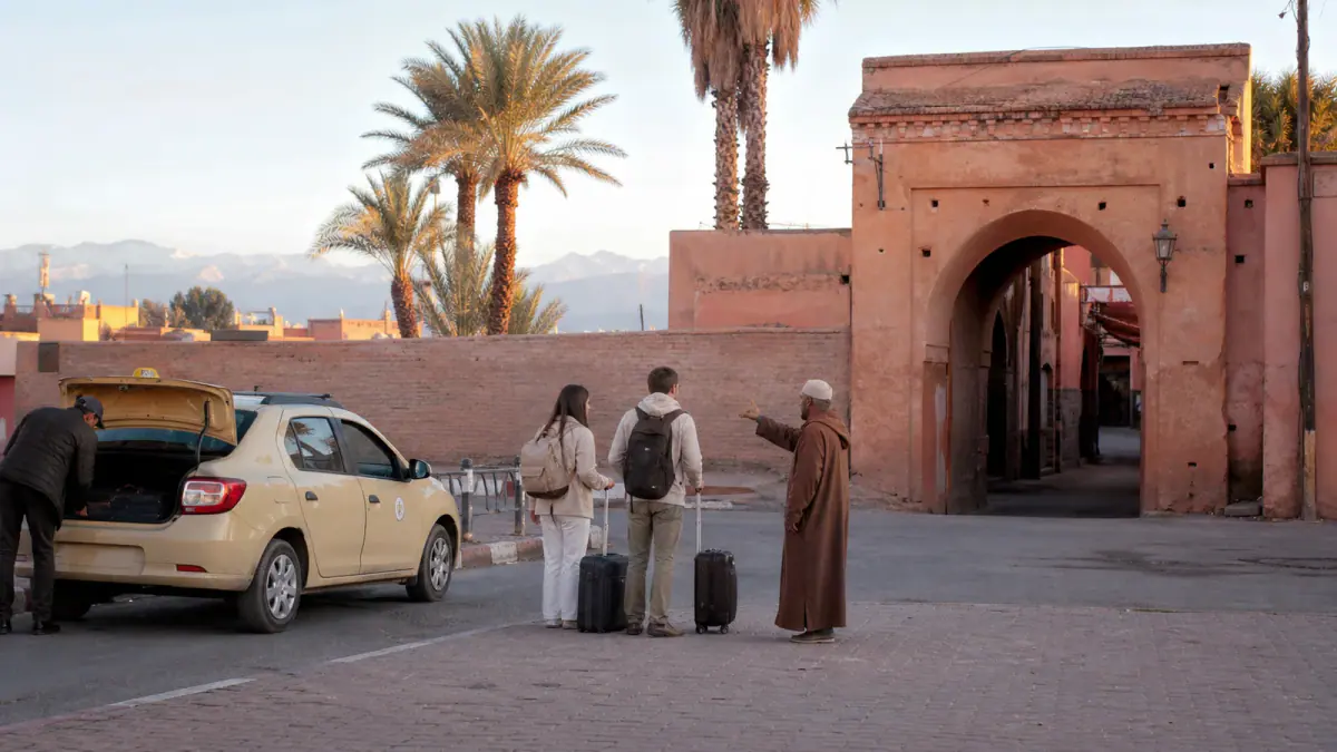 Travelers meeting a porter beside a taxi at the pedestrian edge of Marrakech's ochre medina