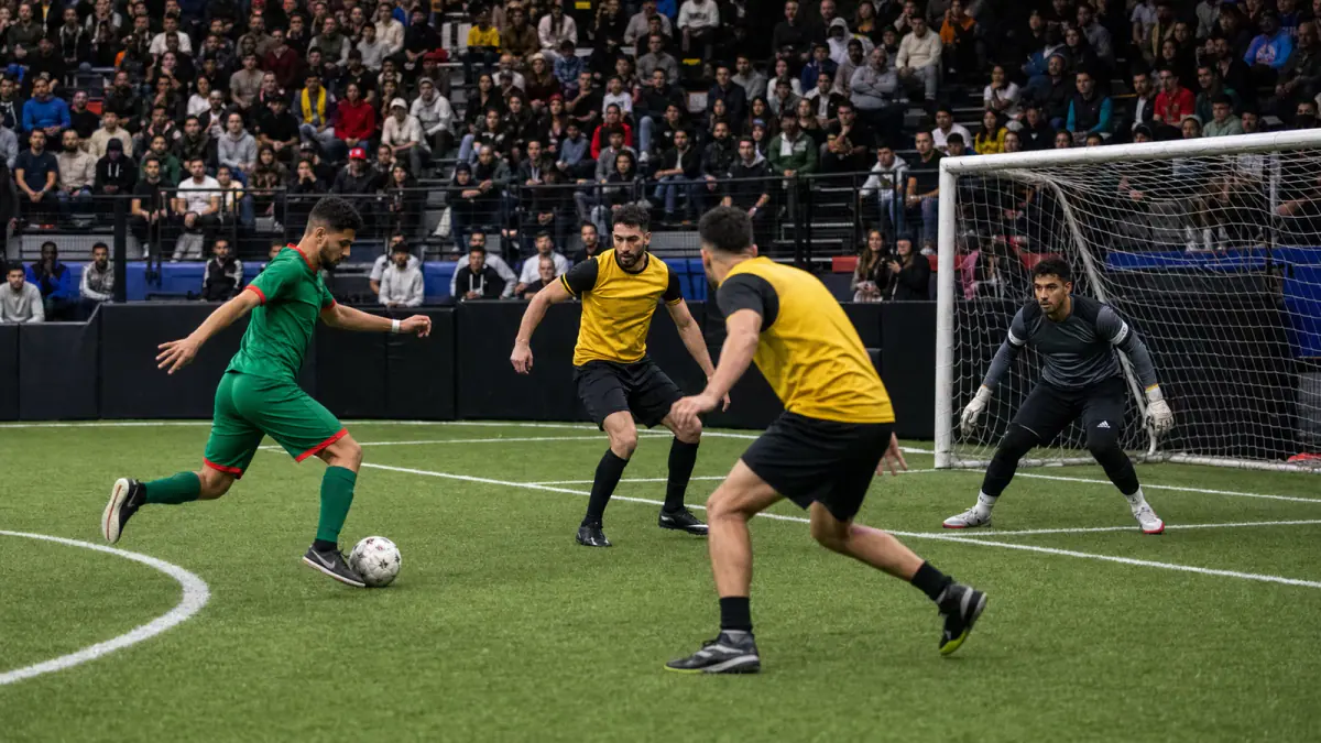Green-kit attacker challenged by yellow-and-black defenders near goal in an indoor arena